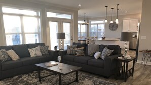 A modern living room with a dark grey sofa, a coffee table, and a fluffy rug. There is a kitchen in the background with white cabinets and stainless steel appliances.