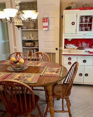 A dining table with a fruit bowl, surrounded by wooden chairs, in a room with a vintage stove and a white door.