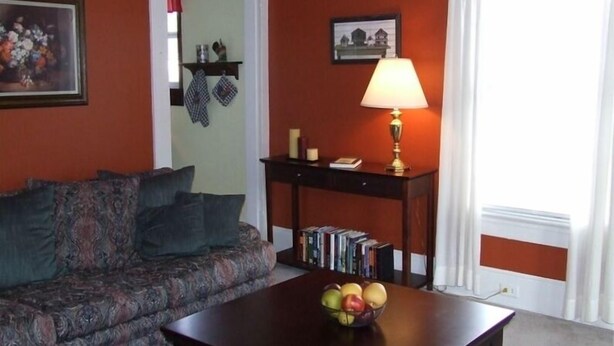 Ein Wohnzimmer mit einer Couch, einem Tisch mit Obstschale, einer Lampe und einem Bücherregal.