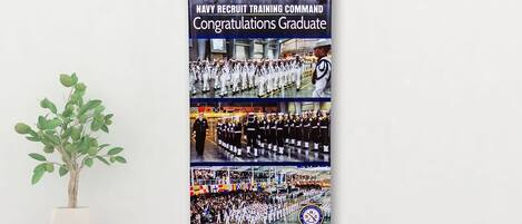 Póster de graduación del Centro de Entrenamiento de Reclutas de la Marina, con una planta blanca en maceta a un lado.