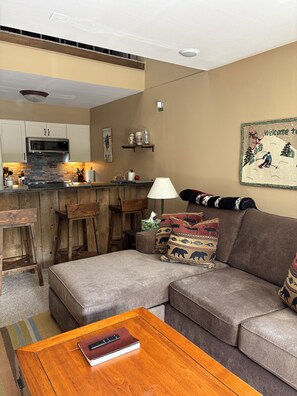 A living room with a sectional sofa, a wooden coffee table, and a kitchen area with a microwave and bar stools.