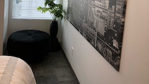 A room with a large black and white cityscape poster on the wall, a window with blinds, a potted plant, and a black ottoman.