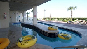 Une piscine dotée de plusieurs bouées gonflables, un espace couvert avec un plafond en béton et une vue sur un jardin aménagé avec des palmiers et des chaises longues.