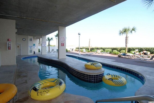 Una piscina con varios flotadores inflables, un área techada con techo de concreto y vistas a un jardín con palmeras y sillas de descanso.