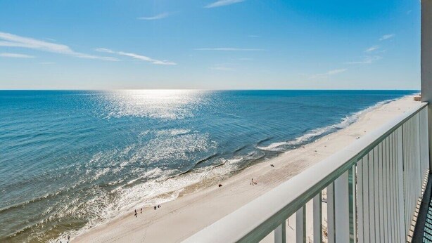 A beach view from a balcony with a railing, clear sky, and sunlight reflecting on the water.