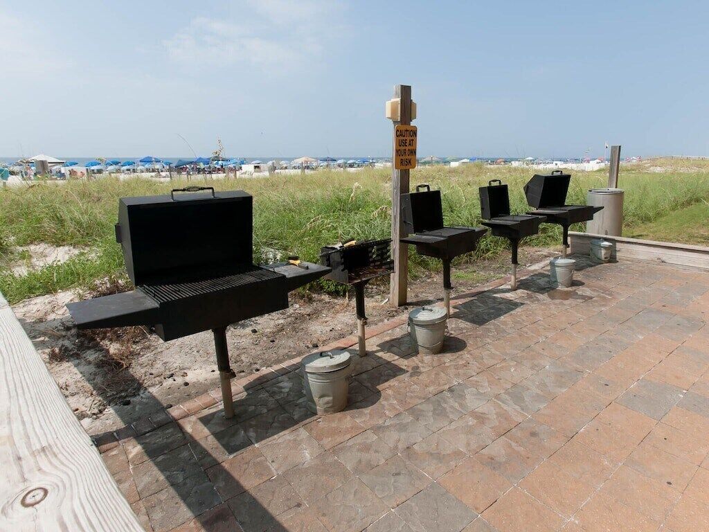 Outdoor barbecue grills and seating on a patio with a warning sign, beach in the background.