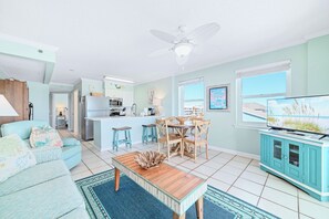 Un salon lumineux avec une cuisine, des meubles de couleur turquoise, un ventilateur de plafond et un téléviseur.
