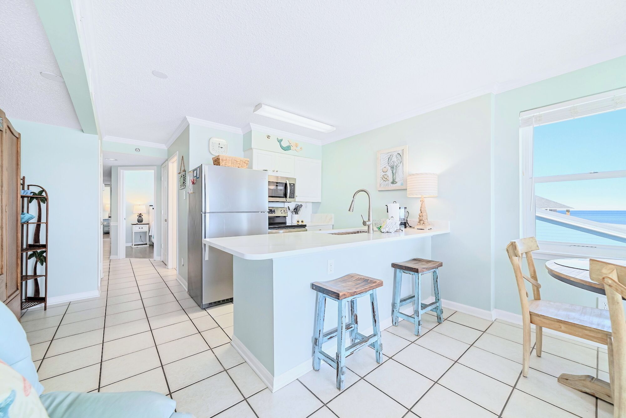Een keuken met een centraal eiland, twee krukken, een koelkast, en uitzicht op de oceaan door een raam.