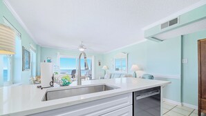 A kitchen with a white countertop, stainless steel sink, and a dishwasher. There is a ceiling fan, a window with a view, and a door leading to another room.