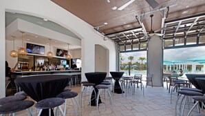 An indoor bar area with high tables and stools, a counter with a bartender, and large windows overlooking a pool area.