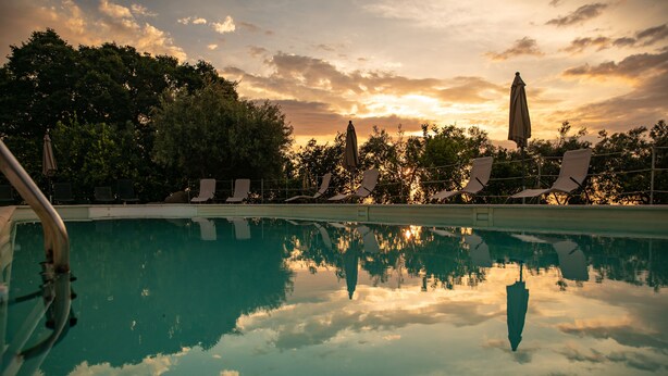 Une piscine entourée d’arbres, avec des chaises longues et des parasols, le tout baigné par la lumière d’un coucher de soleil.