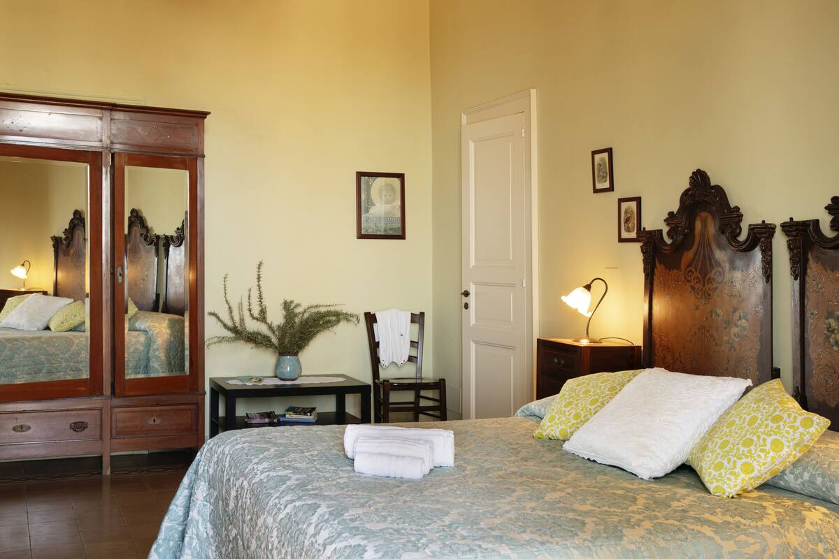 Un dormitorio con un espejo grande, una cama con ropa de cama estampada, una silla, una mesita con una lámpara y una puerta.
