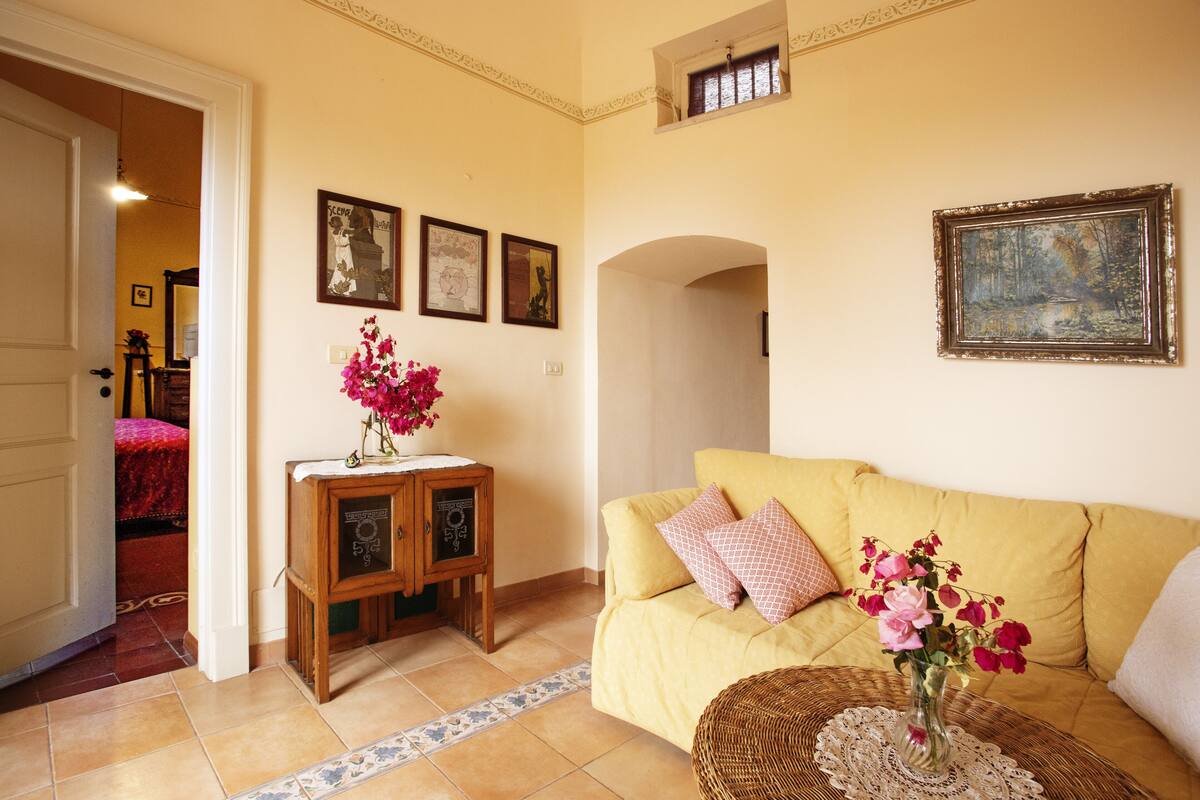 Un salón con un sofá amarillo, un pequeño armario de madera con un jarrón de flores y cuadros enmarcados en la pared.