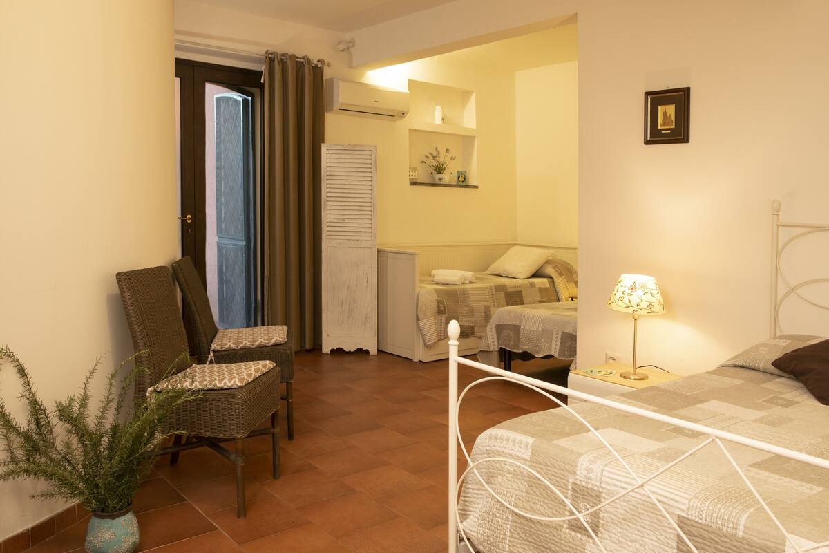 Un dormitorio con cama, una silla, mesita de noche con lámpara, estante de pared con objetos decorativos y una puerta que da a otra habitación.