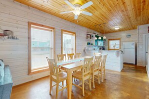 A wooden-paneled room with a dining table and chairs, a ceiling fan, and a kitchen area with a refrigerator and sink.