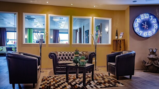 A lobby with a sofa, armchairs, a coffee table, a large wall clock, and a checkered rug. - Hotel Aulmann (Trier)