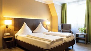 A hotel room with a large bed, two bedside lamps, a chair, a small table, and a window with curtains. - Hotel Aulmann (Trier)