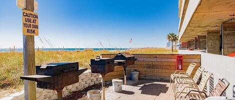 Une terrasse extérieure équipée de barbecues, agrémentée d’un panneau d’avertissement et offrant une vue sur la plage.