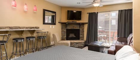 A hotel room with a bed, a fireplace, a TV, a balcony with a view, and three bar stools.