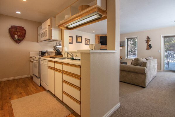A modern kitchen with white appliances, wooden cabinets, and a central island. There is a dining area with a sofa and a large window. - RMR: Balsam Lodge B4 Condo in The Aspens/Wilson, WY (Wilson)