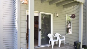 Een overdekte veranda met twee witte plastic stoelen, een zwarte prullenbak en een schuifdeur die naar de binnenruimte leidt.