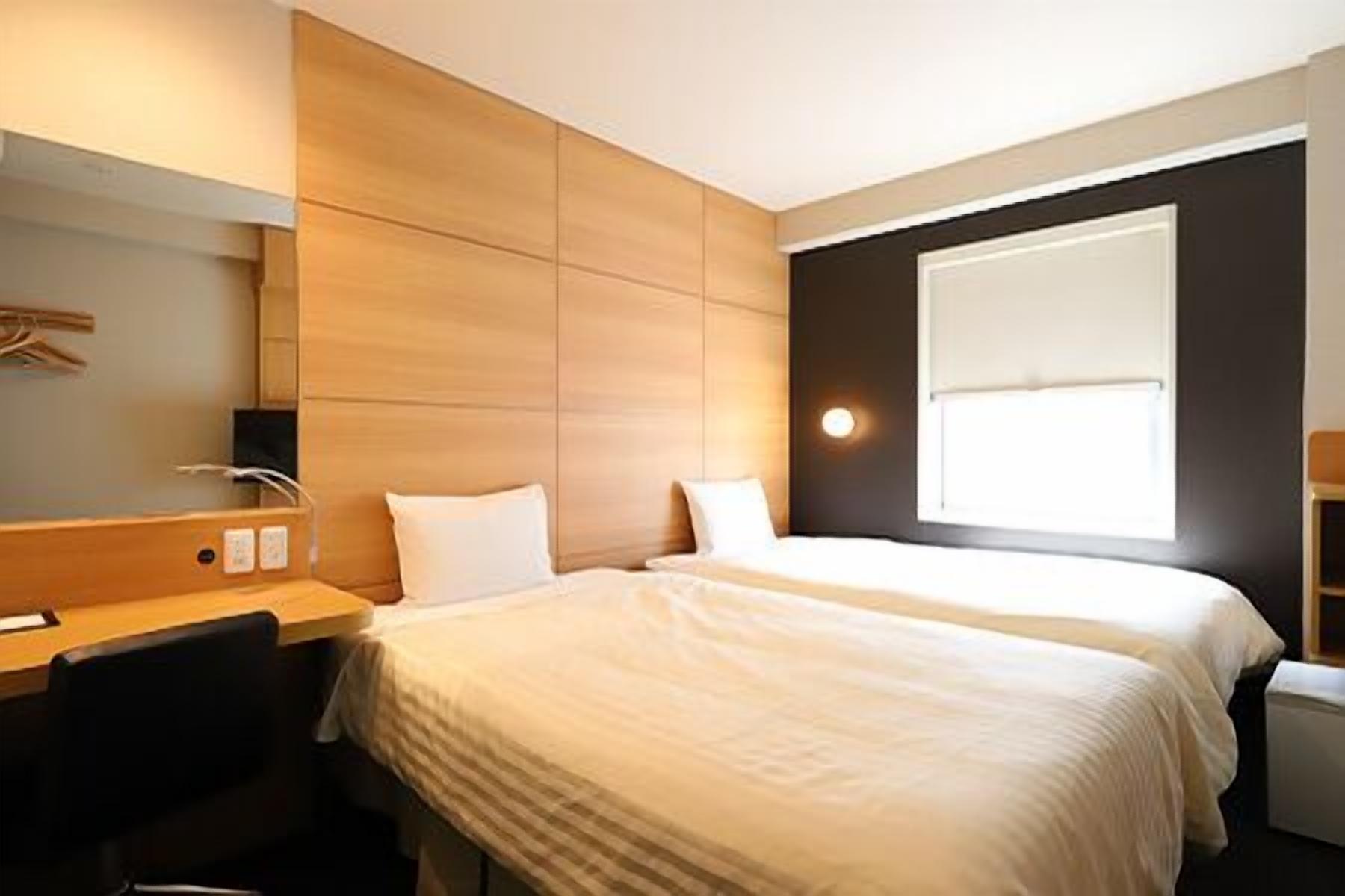 Ein Hotelzimmer mit zwei Betten, einem Schreibtisch, einem Stuhl, einem Spiegel und einem Fenster.