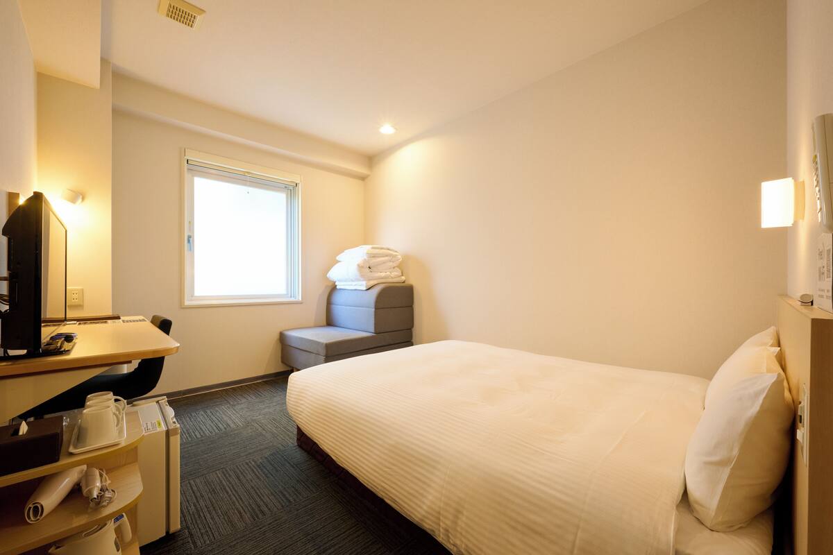 Een hotelkamer met een bed, een bureau met een computer, een stoel, een raam en een wandairconditioner.
