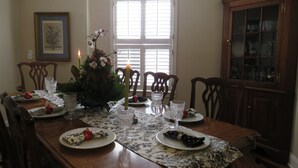 A dining room with a wooden table set for four, surrounded by chairs. There is a window with blinds, a framed picture on the wall, and a cabinet with glass doors.
