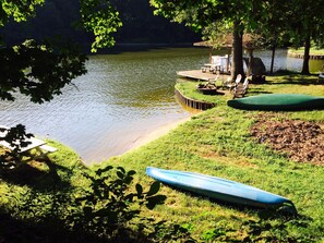 Terraza junto al lago con un bote, una zona de estar cubierta y una orilla con césped.