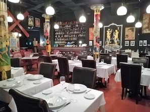 Interior de restaurante com mesas postas para refeições, área de bar ao fundo e diversas obras de arte nas paredes.