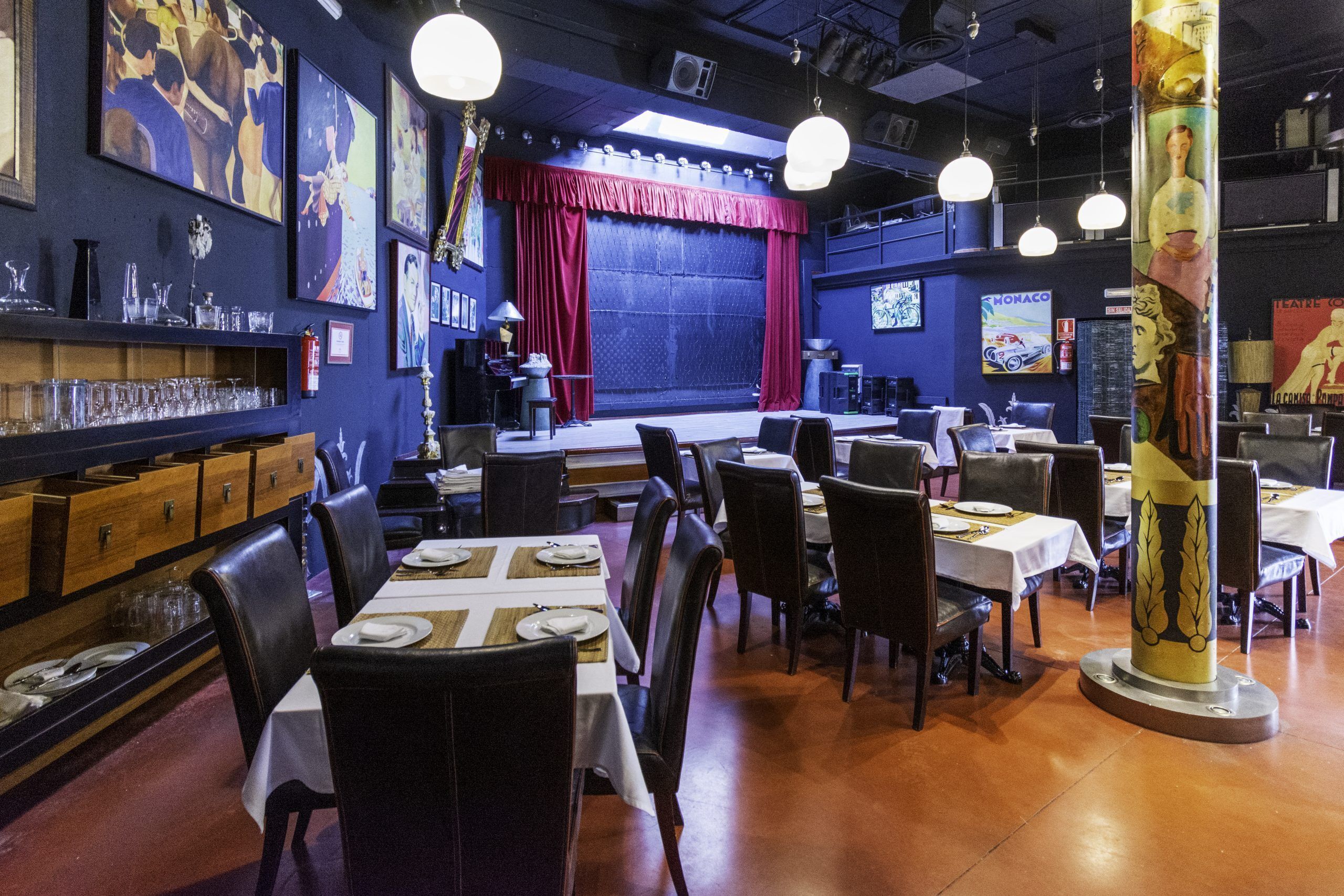 Restaurante com mesas postas para refeições, palco com cortinas e obras de arte nas paredes.