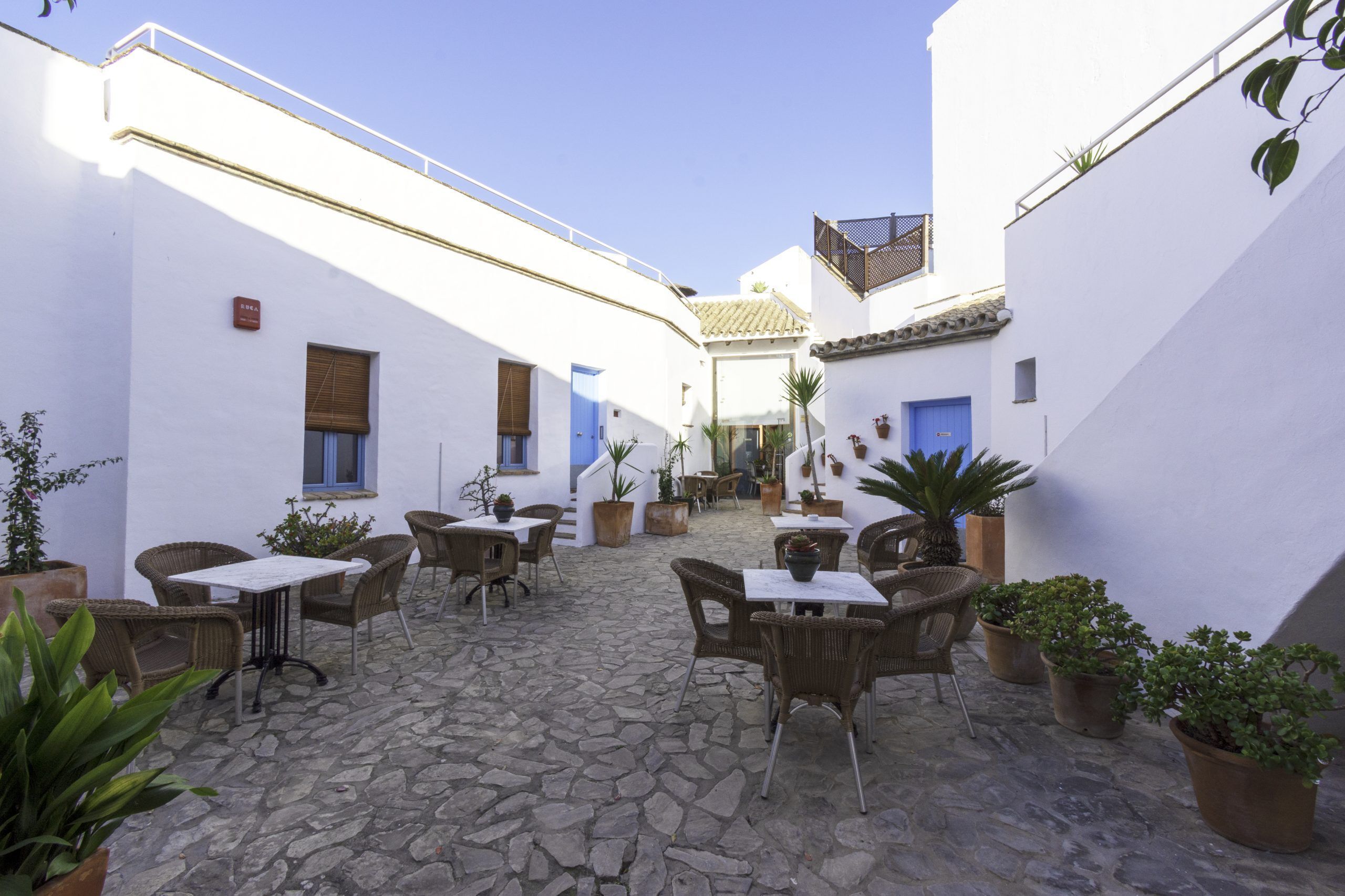 Un patio con suelo de piedra, paredes blancas, puertas azules, mobiliario de mimbre y plantas en macetas.