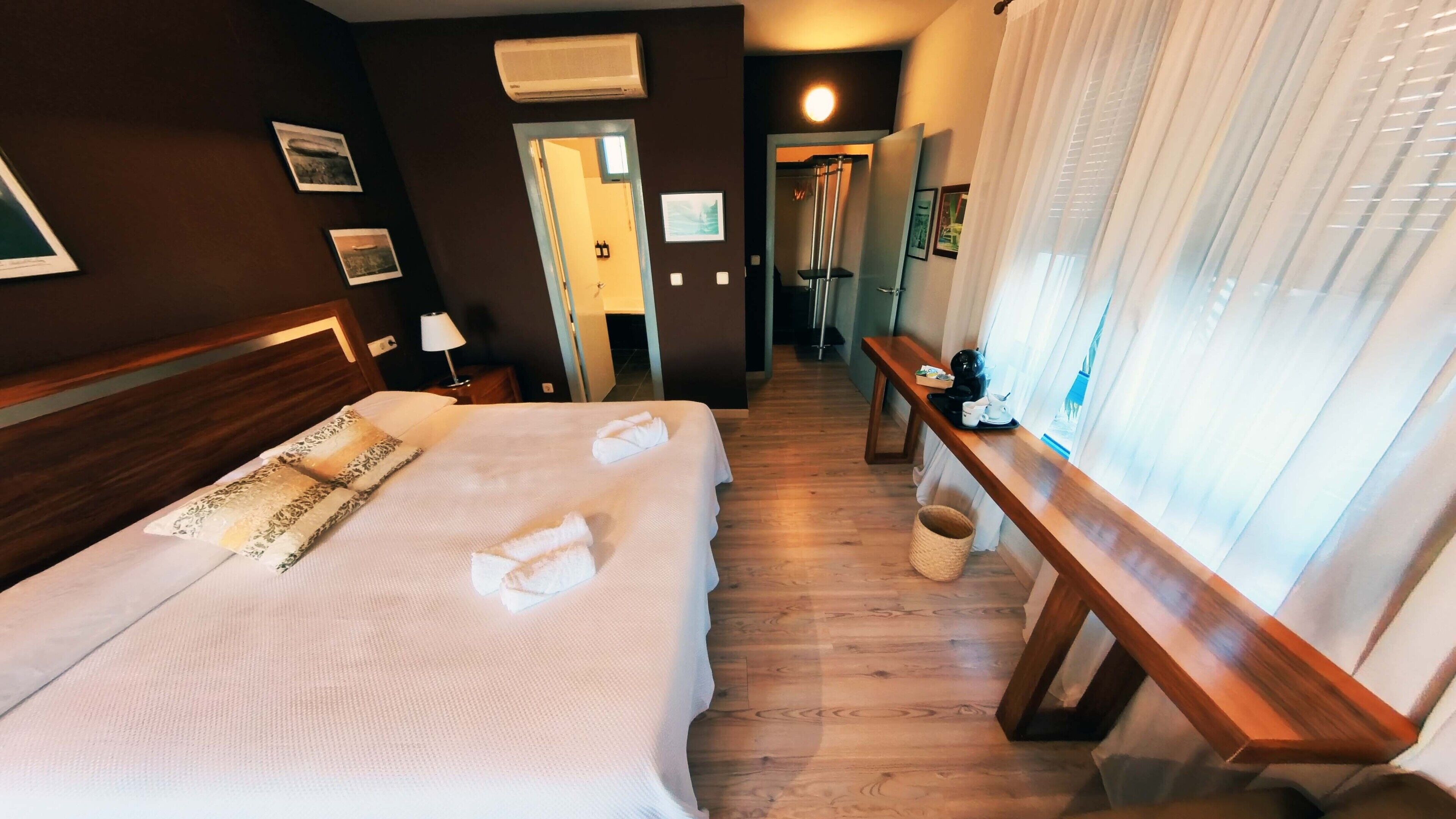 A hotel room with a large bed, a wooden bench, a window with curtains, and a hallway leading to a bathroom.