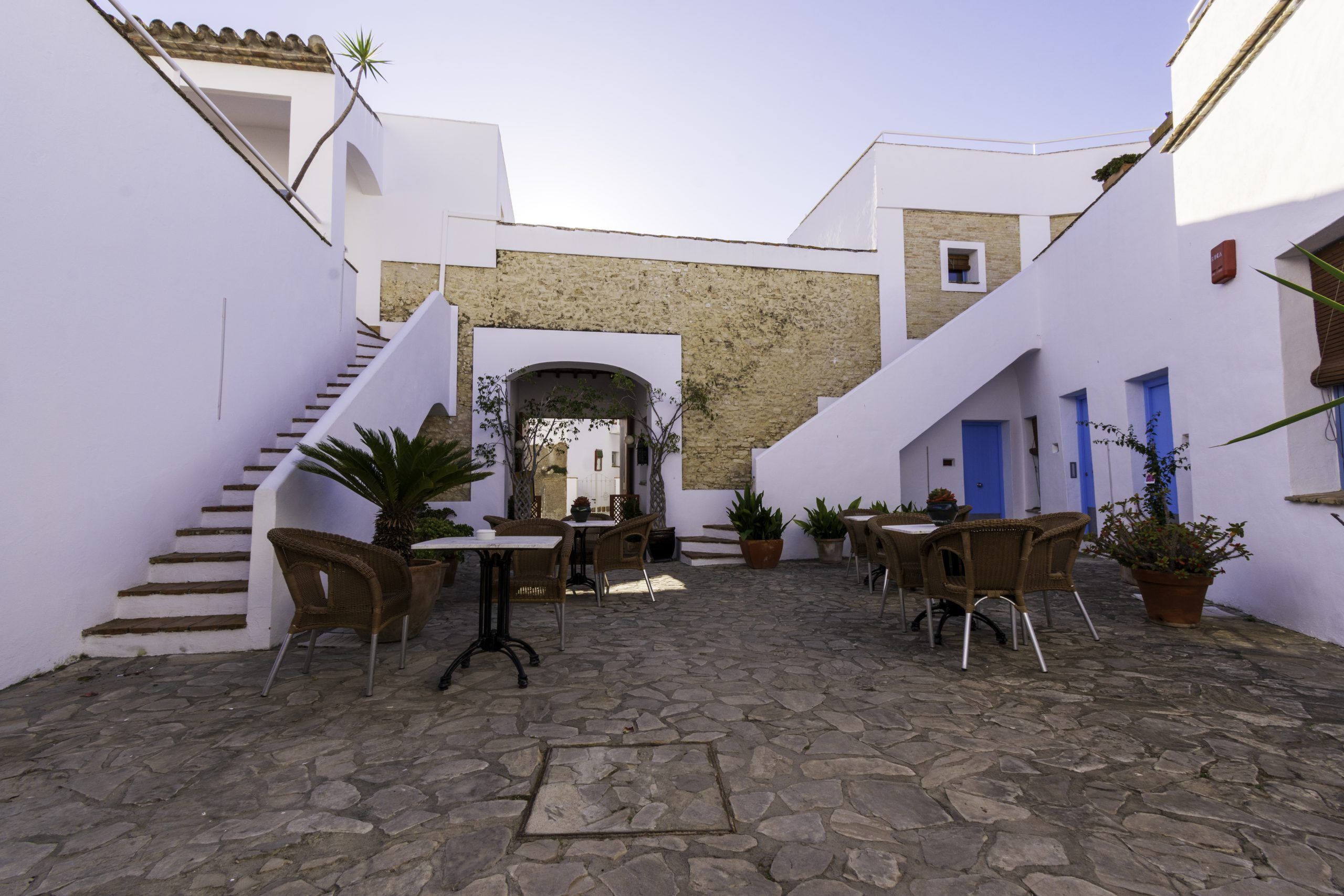 Un patio con suelo de piedra, paredes blancas y puertas azules. Hay sillas de mimbre y plantas en macetas.