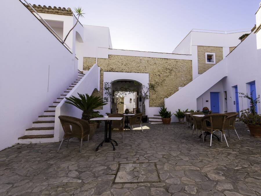 Un patio con suelo de piedra, paredes blancas y puertas azules. Hay sillas de mimbre y plantas en macetas.