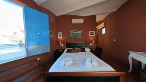 Habitación de hotel con una cama, dos mesitas de noche, un ventanal con puerta azul y un cuadro de paisaje urbano en la pared.
