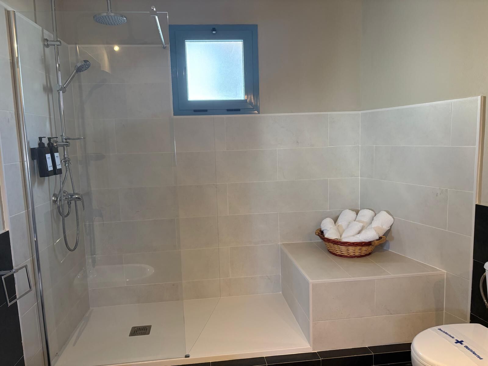 Una ducha con puerta de cristal, un ventanal pequeño y una cesta de toallas sobre un pedestal blanco.