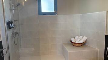 Una ducha con puerta de cristal, un ventanal pequeño y una cesta de toallas sobre un pedestal blanco.