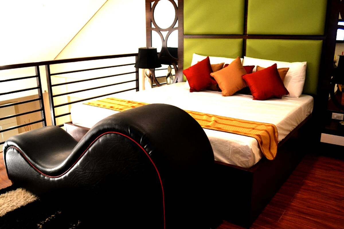 Een slaapkamer met een bed, kussens, een hoofdbord, een nachtkastje en een lamp.