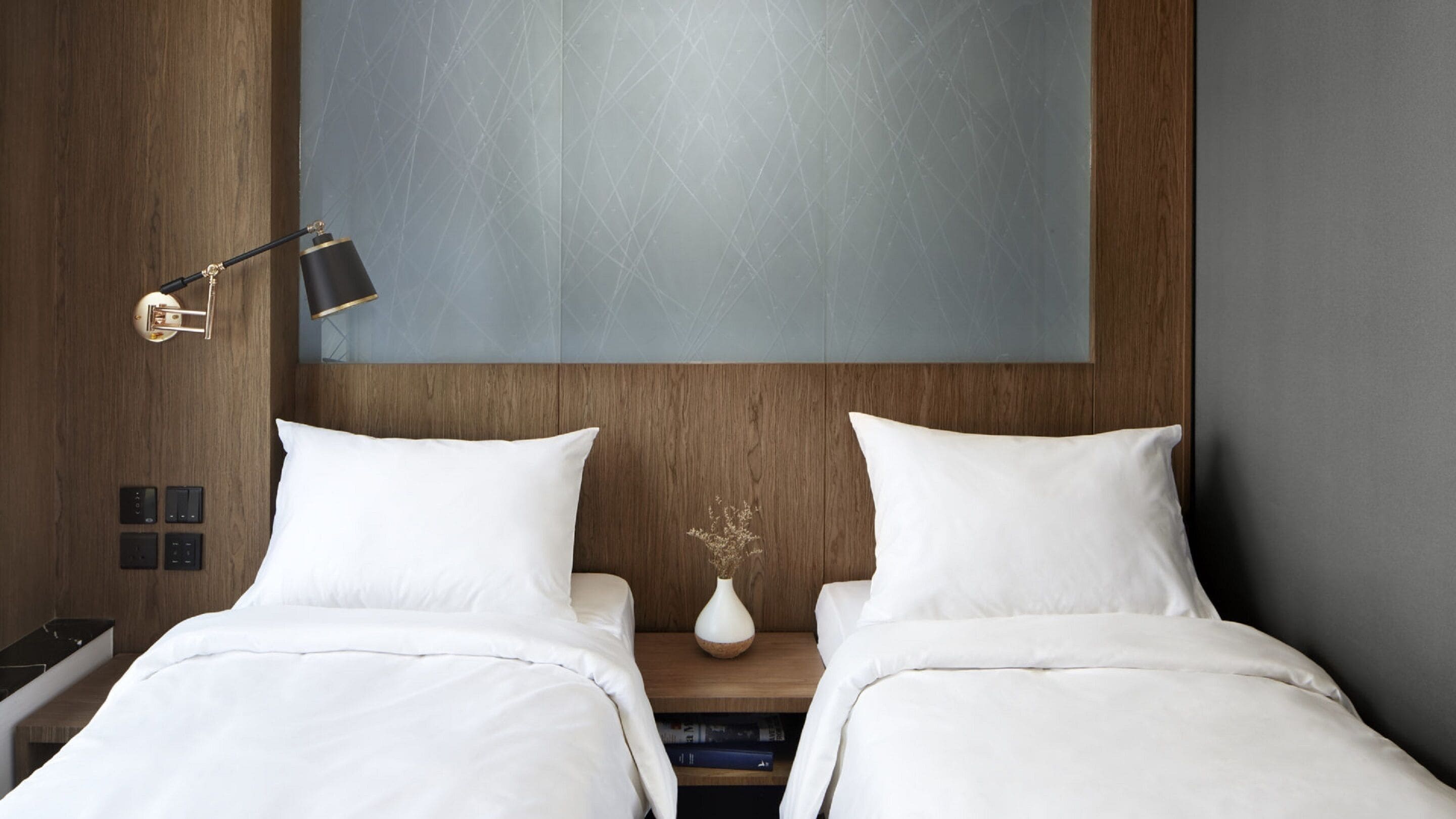 Une chambre d’hôtel avec une tête de lit en bois, deux lits blancs, une table de chevet avec un vase et une lampe fixée au mur.