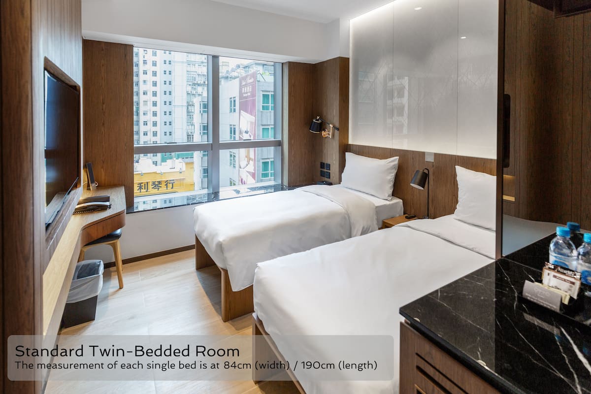 A standard twin-bedded room with two beds, a desk, a chair, a TV, and a large window offering a city view.