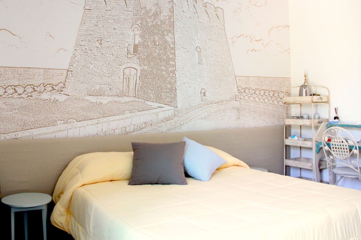 Quarto com cama, almofadas, uma mesinha redonda e um mural na parede com um castelo.