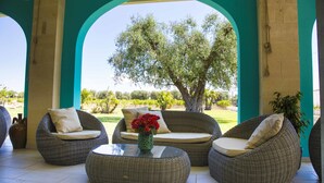 A covered outdoor seating area with wicker furniture and a glass table, overlooking a garden with a large tree. - Tenuta Donna Caterina (Mesagne)