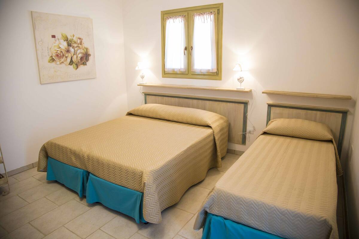 Quarto com duas camas, uma janela com cortinas, mesa de cabeceira com abajur e um quadro na parede.