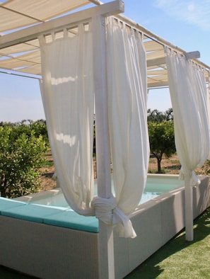 Un área de estar al aire libre techada con cortinas blancas atadas en las esquinas, una piscina y vistas a la vegetación.
