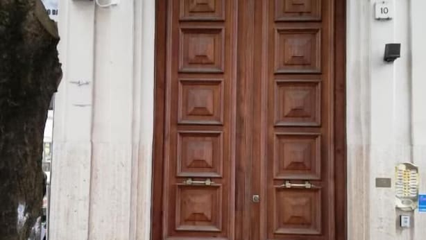 A double wooden door with a brass doorknob, a blue mat at the entrance, and a small plaque on the wall. - Landlord in Trastevere (Rome)
