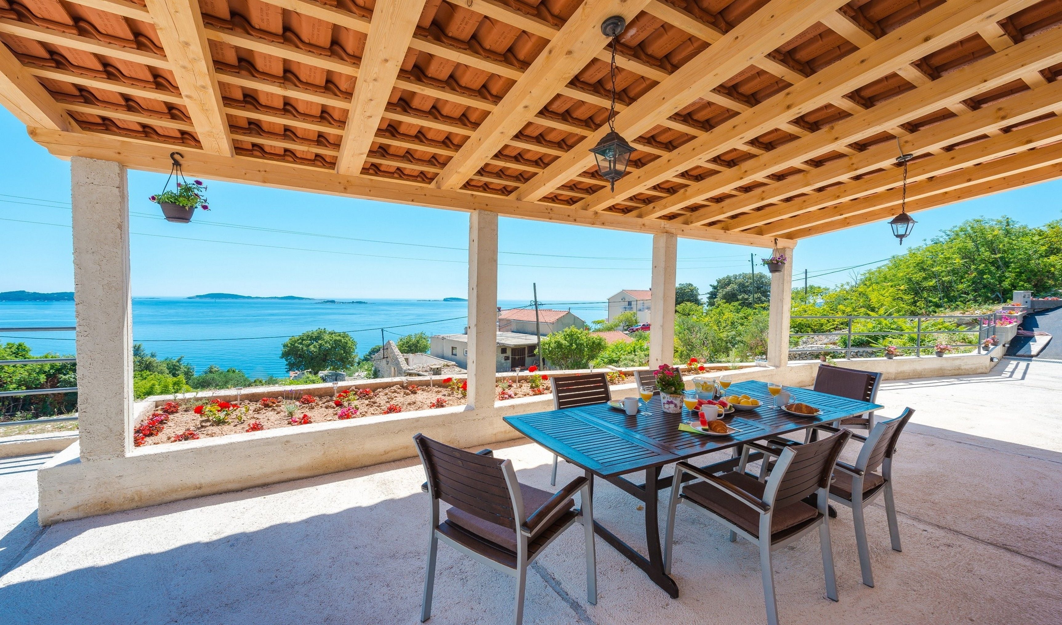 A covered outdoor seating area with a table and chairs, overlooking a body of water and a coastal landscape.