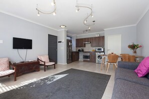 Ett modernt vardagsrum med en grå soffa, ett soffbord i trä och en platt-TV. Här finns också en köksdel med träskåp och ett matbord med stolar.