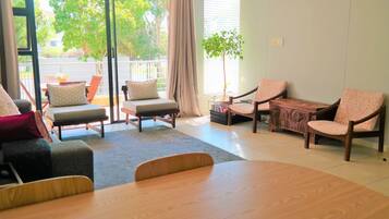A spacious living area with a large sliding glass door leading to a balcony, a wooden dining table, and two armchairs with patterned cushions.
