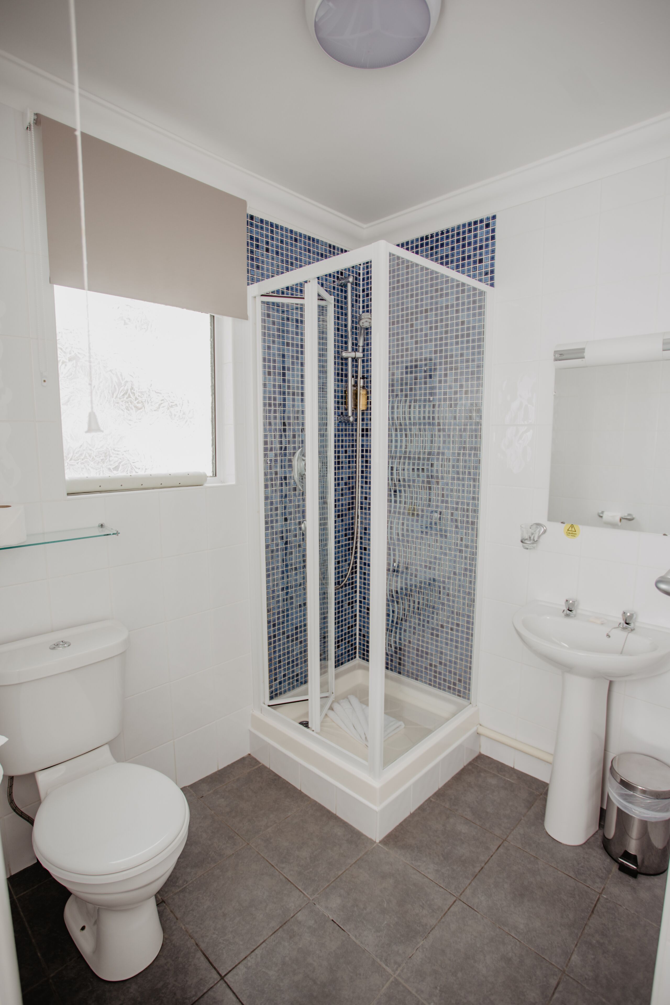 A bathroom with a white toilet, a glass shower enclosure with blue mosaic tiles, a white sink, and a window with a hanging light fixture.